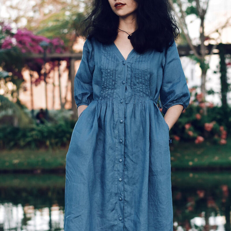 A blue 100% linen midi dress with a soft V-neckline and fabric-covered buttons down the front. Designed with delicate pintuck pleats on the bodice and puffy 3/4 sleeves, this dress blends romantic vintage charm with breathable natural fabric. The gathered waist adds gentle structure for a relaxed yet flattering silhouette.