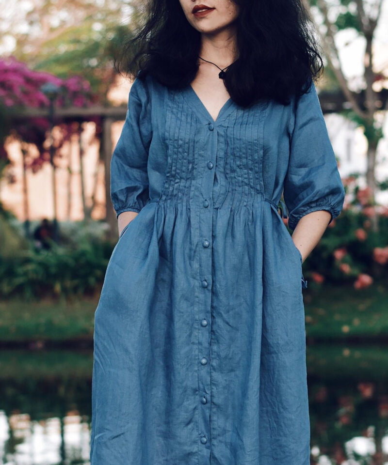 A blue 100% linen midi dress with a soft V-neckline and fabric-covered buttons down the front. Designed with delicate pintuck pleats on the bodice and puffy 3/4 sleeves, this dress blends romantic vintage charm with breathable natural fabric. The gathered waist adds gentle structure for a relaxed yet flattering silhouette.