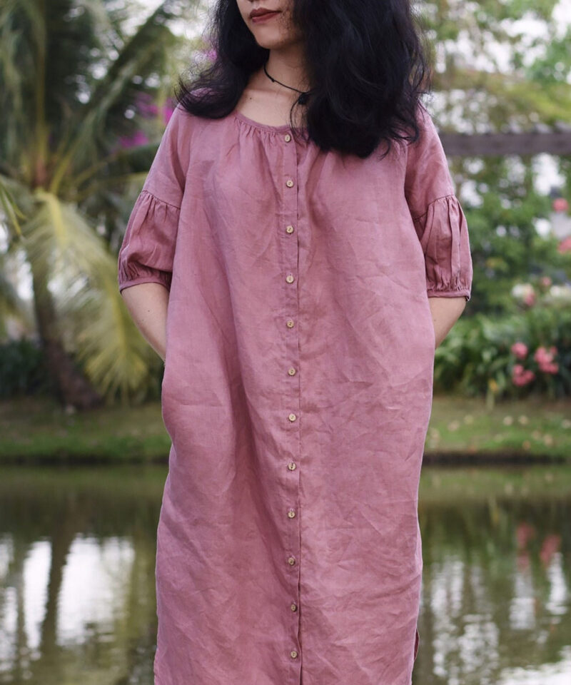 Woman wearing a loose-fitting midi dress in dusty rose pink, crafted from 100% natural linen. The dress features a button-down front and gathered half sleeves, offering a breezy and effortless silhouette perfect for warm weather. Captured in a lush outdoor garden, this design blends comfort, elegance, and organic charm.