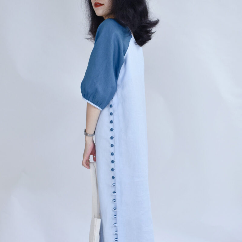 A closer side view reveals the clean tailoring and elegant side button placket of this two-tone linen dress. The deep blue sleeve detail adds structure to the lightweight ivory body, while the line of fabric buttons emphasizes vertical balance. Its modern minimalist cut blends comfort and natural charm for everyday wear.