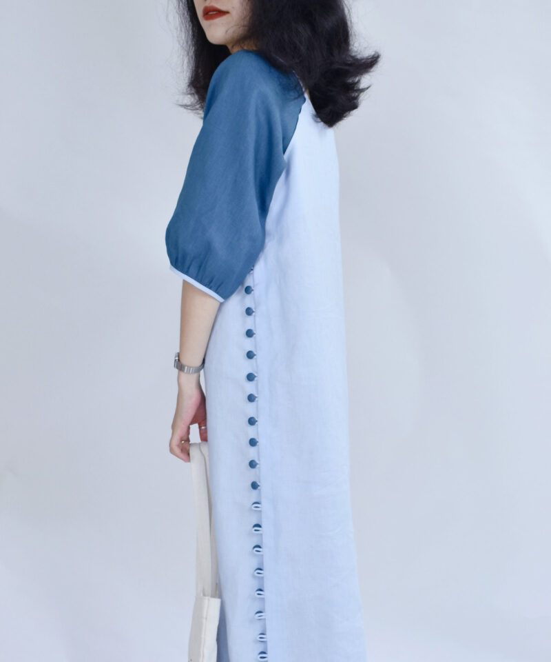 A closer side view reveals the clean tailoring and elegant side button placket of this two-tone linen dress. The deep blue sleeve detail adds structure to the lightweight ivory body, while the line of fabric buttons emphasizes vertical balance. Its modern minimalist cut blends comfort and natural charm for everyday wear.