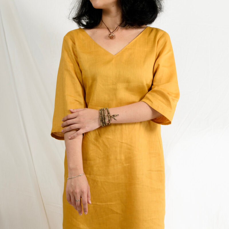 A minimalist midi linen dress in mustard yellow, crafted from 100% natural linen with a soft, breathable texture. The dress features a clean V-neckline, relaxed half sleeves, and a straight silhouette that drapes naturally. Perfect for slow fashion lovers seeking elegance and comfort for everyday wear or warm-weather outings.