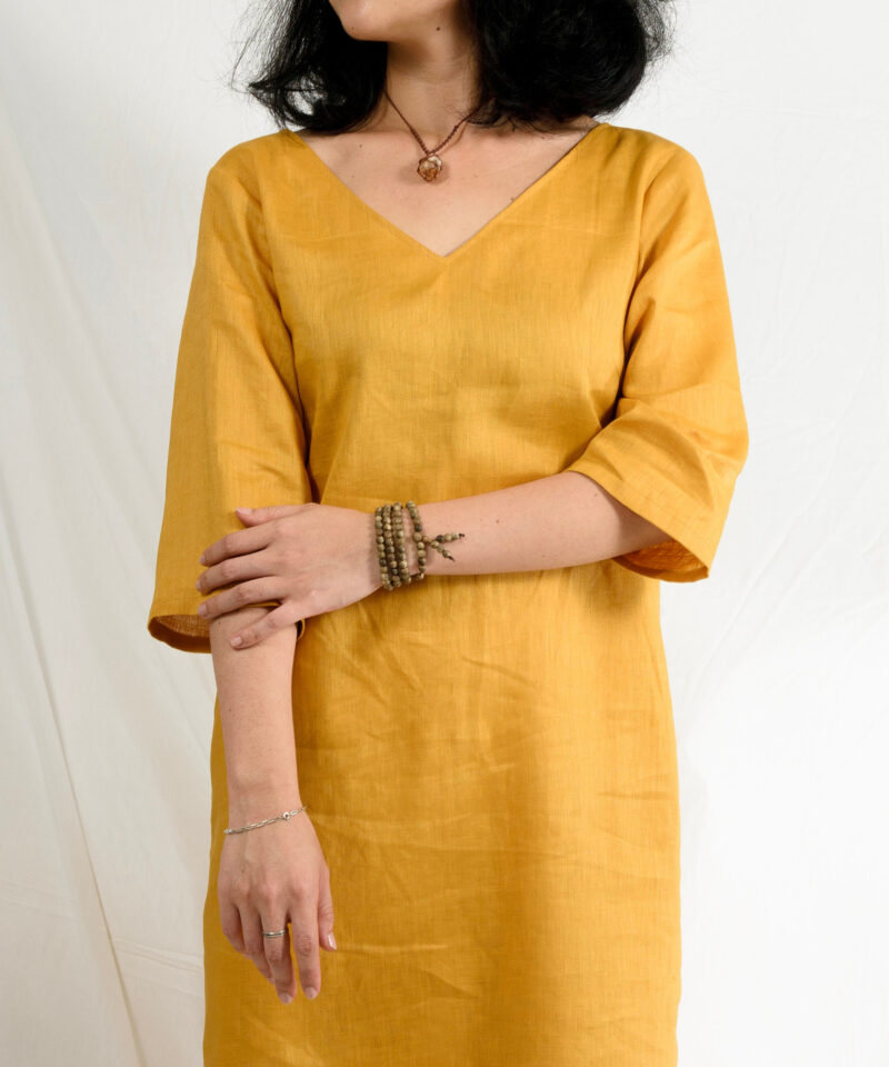 A minimalist midi linen dress in mustard yellow, crafted from 100% natural linen with a soft, breathable texture. The dress features a clean V-neckline, relaxed half sleeves, and a straight silhouette that drapes naturally. Perfect for slow fashion lovers seeking elegance and comfort for everyday wear or warm-weather outings.
