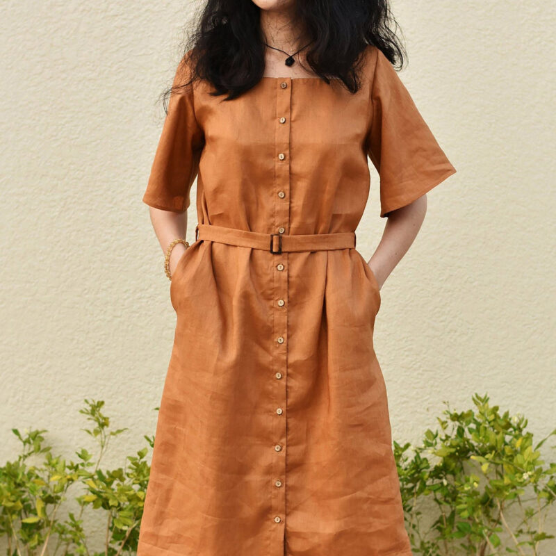 This earthy brown linen dress features a structured silhouette with a matching fabric belt that accentuates the waist. Crafted from breathable 100% natural linen, it offers a clean, tailored front with a full button-down design and discreet side pockets. A minimal yet confident style for warm weather days.