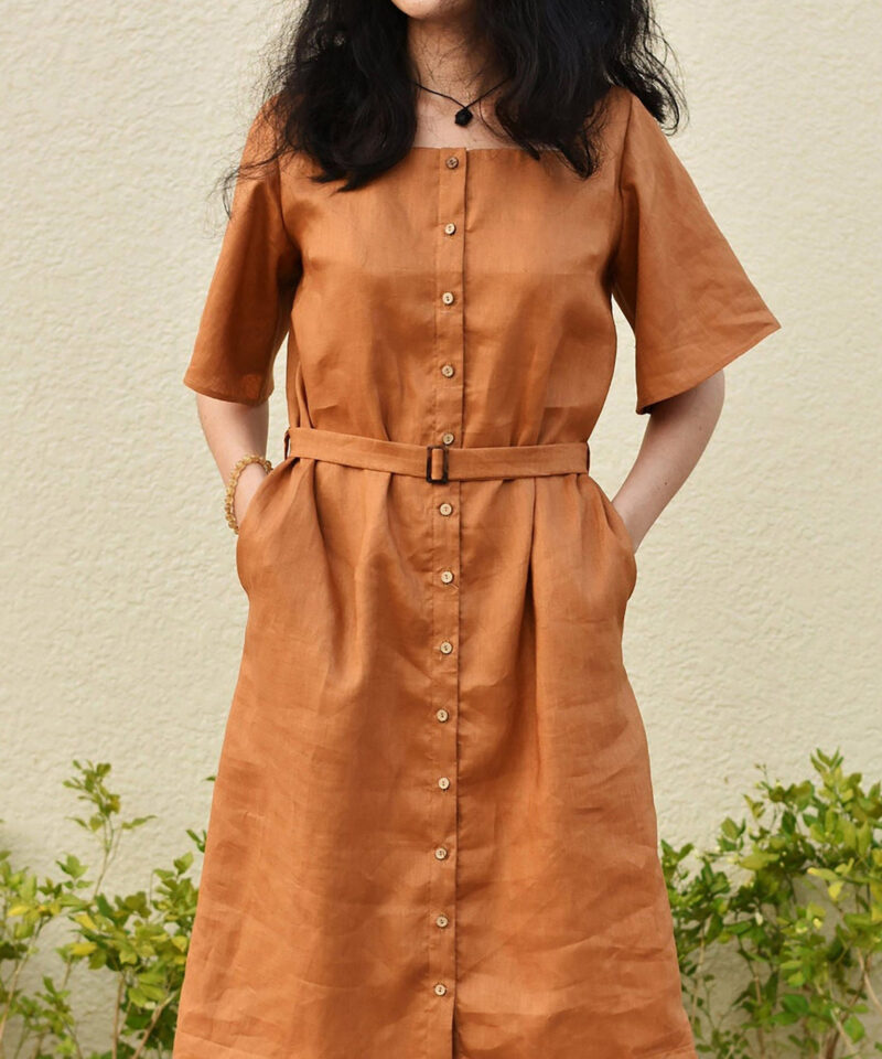 This earthy brown linen dress features a structured silhouette with a matching fabric belt that accentuates the waist. Crafted from breathable 100% natural linen, it offers a clean, tailored front with a full button-down design and discreet side pockets. A minimal yet confident style for warm weather days.