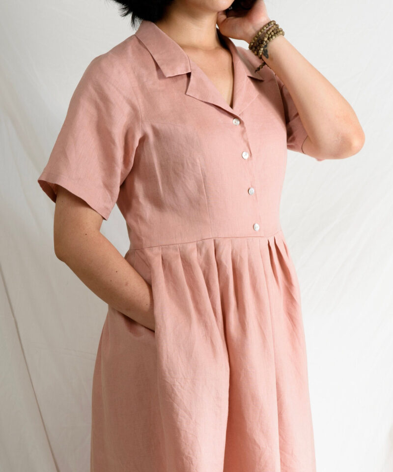 Crafted from breathable 100% linen, this pastel pink dress features a relaxed fit with short sleeves, a soft pleated skirt, and delicate mother-of-pearl buttons. The natural texture of linen pairs beautifully with the structured collar, offering a timeless look inspired by effortless summer dressing.