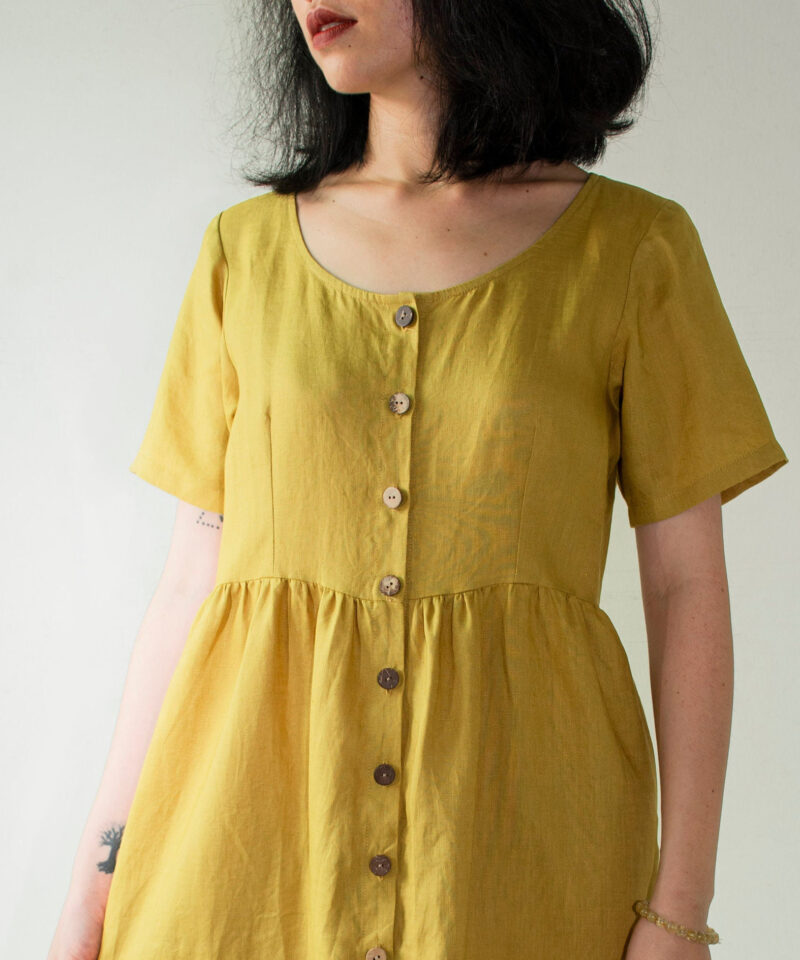 Soft yellow linen midi dress made from 100% natural fabric, featuring a round neckline, short sleeves, and a gently gathered waist. Wooden buttons run the full length of the front, adding a rustic charm and vintage-inspired touch. A breathable, lightweight choice for minimalist summer styling.