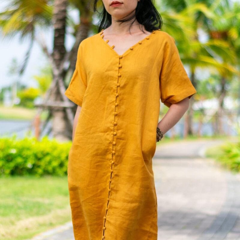 A mustard yellow linen dress with a soft V-neckline and handcrafted fabric-covered buttons running down the center. Made from breathable natural linen, the dress features a straight, relaxed silhouette and rolled short sleeves, ideal for summer outings or casual strolls. Inspired by tropical sunshine and slow living aesthetics.