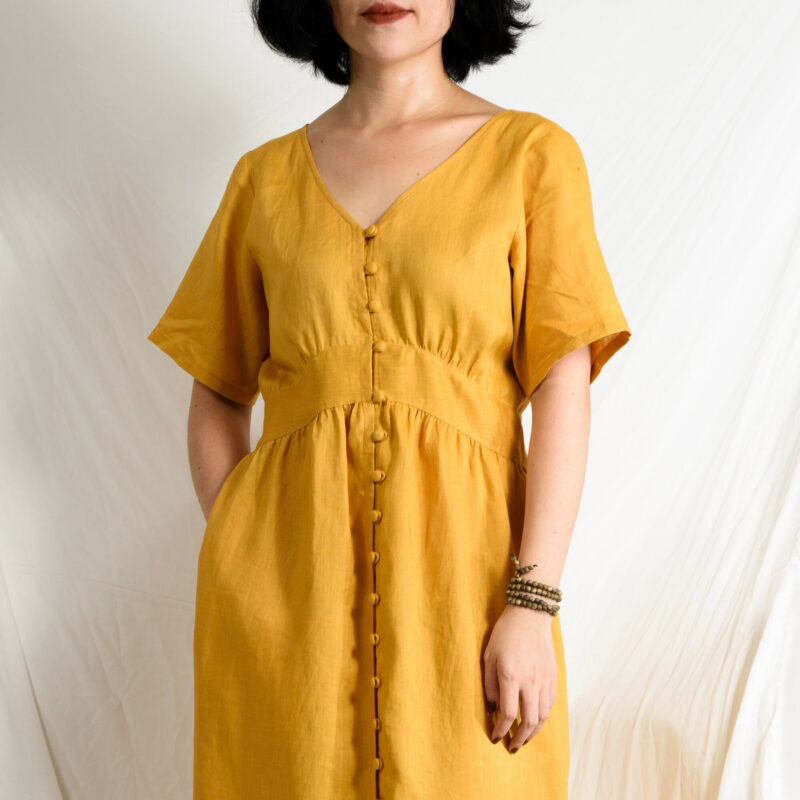 Crafted from 100% natural linen, this golden yellow dress features a flattering V-neckline and a soft empire waist that gently accentuates the figure. The covered button closure adds a handcrafted charm, while short sleeves and midi length offer effortless elegance. A breezy and timeless piece for warm, sunlit days.