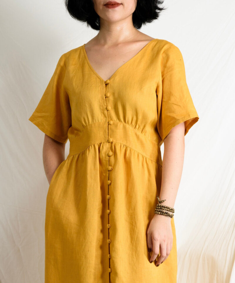 Crafted from 100% natural linen, this golden yellow dress features a flattering V-neckline and a soft empire waist that gently accentuates the figure. The covered button closure adds a handcrafted charm, while short sleeves and midi length offer effortless elegance. A breezy and timeless piece for warm, sunlit days.
