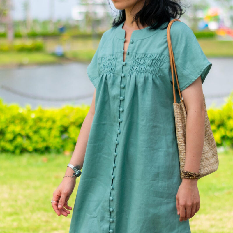 Woman walking in a soft green linen dress on a sunlit path. The dress features fabric-covered buttons from top to bottom and gentle gathers around the chest. Crafted from breathable natural linen, the flowy silhouette and simple elegance reflect a slow fashion lifestyle ideal for warm weather days.
