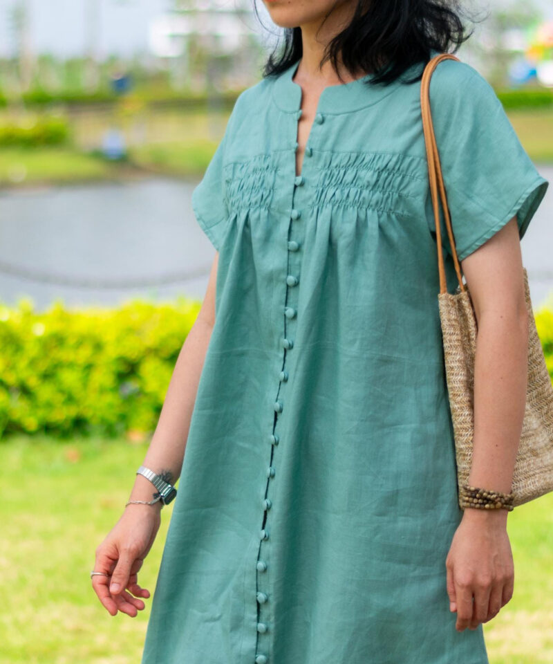 Woman walking in a soft green linen dress on a sunlit path. The dress features fabric-covered buttons from top to bottom and gentle gathers around the chest. Crafted from breathable natural linen, the flowy silhouette and simple elegance reflect a slow fashion lifestyle ideal for warm weather days.