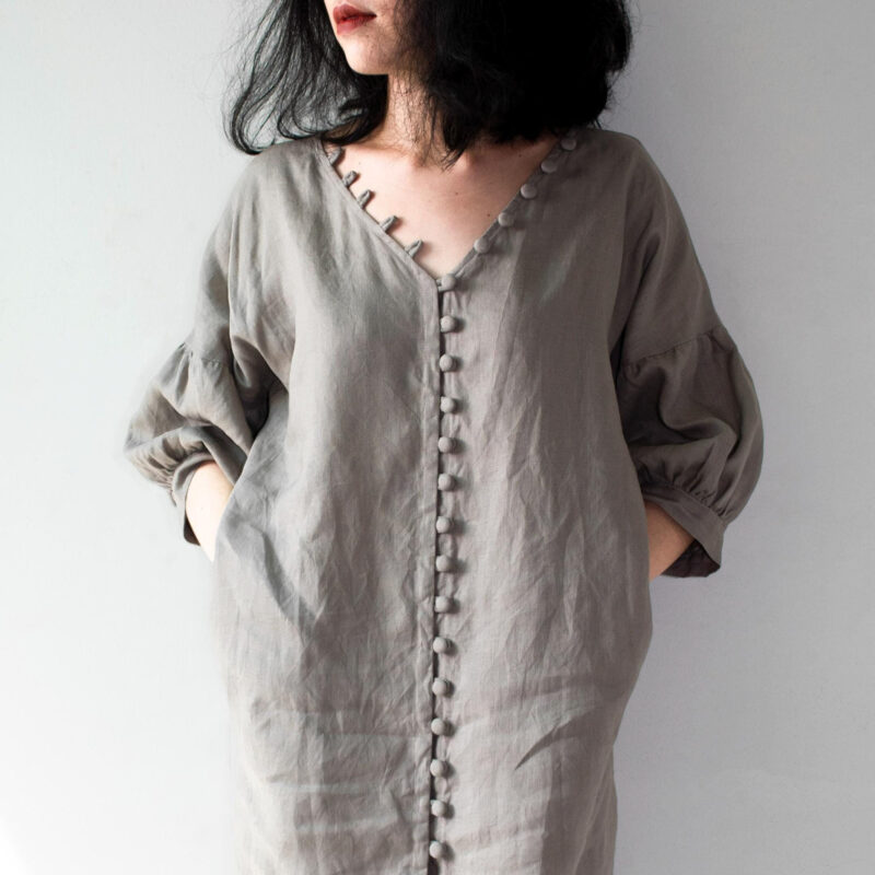 A handmade grey linen dress with a deep V-neckline, featuring a row of fabric-covered buttons running vertically down the center. The 100% natural linen fabric offers a soft drape and breathable comfort, ideal for warm seasons. Designed with roomy balloon sleeves, this dress embraces minimalist aesthetics with earthy tones and effortless elegance.