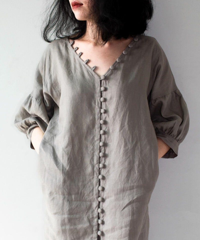 A handmade grey linen dress with a deep V-neckline, featuring a row of fabric-covered buttons running vertically down the center. The 100% natural linen fabric offers a soft drape and breathable comfort, ideal for warm seasons. Designed with roomy balloon sleeves, this dress embraces minimalist aesthetics with earthy tones and effortless elegance.