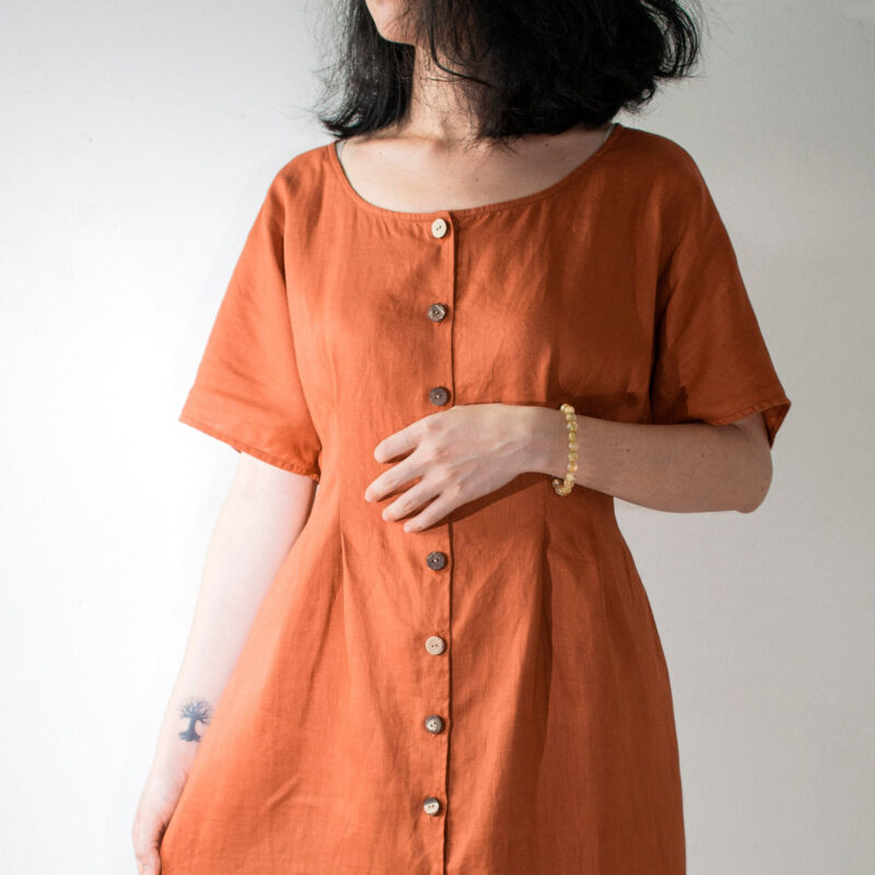 Crafted from breathable 100% linen, this rust-toned dress embraces a relaxed yet flattering silhouette. The wide scoop neckline and short sleeves offer effortless charm, while the handmade wooden buttons add a rustic touch. Ideal for warm days and minimal styling, this dress blends comfort with timeless aesthetics.