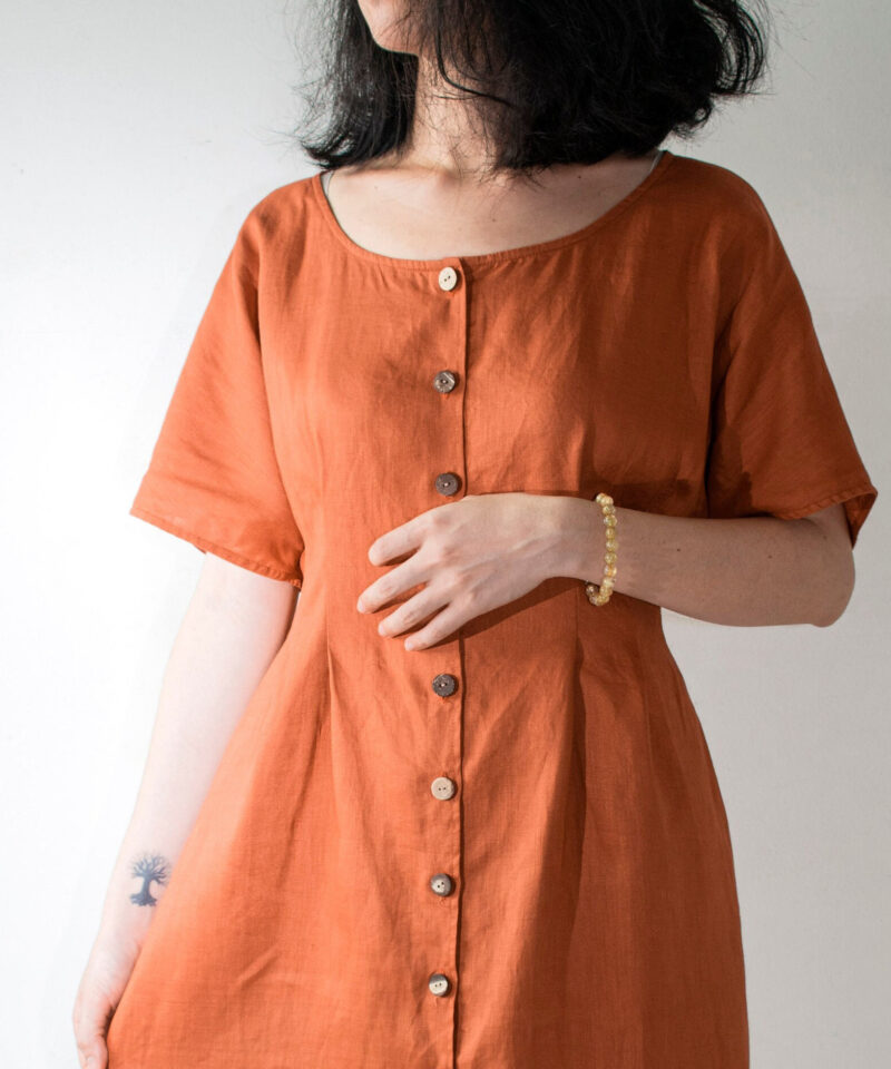 Crafted from breathable 100% linen, this rust-toned dress embraces a relaxed yet flattering silhouette. The wide scoop neckline and short sleeves offer effortless charm, while the handmade wooden buttons add a rustic touch. Ideal for warm days and minimal styling, this dress blends comfort with timeless aesthetics.