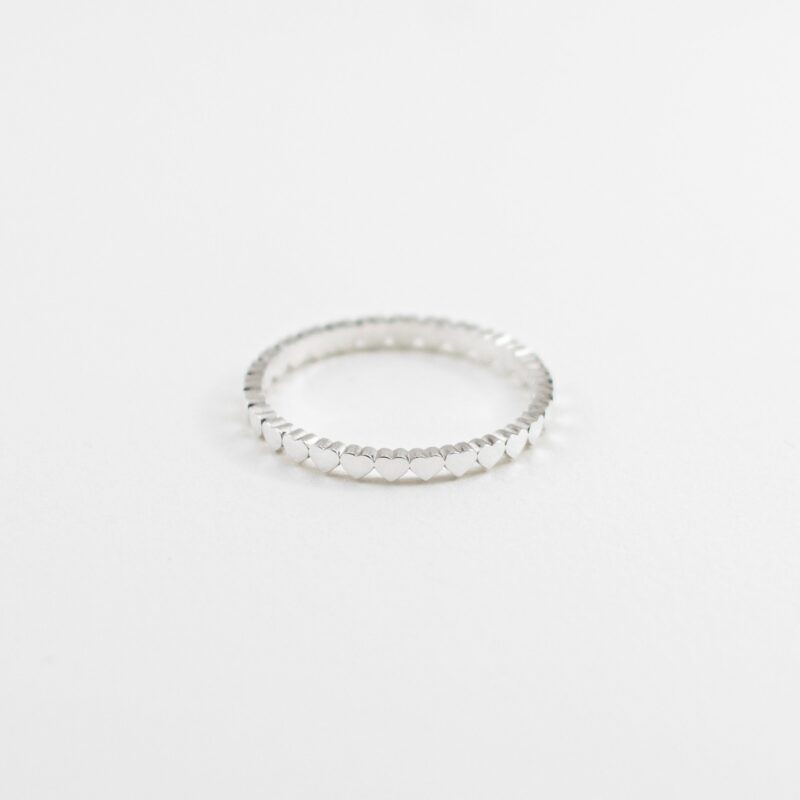 A minimalist sterling silver ring featuring a delicate continuous band of tiny sculpted hearts, photographed from a soft angle with a shallow depth of field on a clean white background. This romantic and symbolic design captures subtle texture and polished elegance, perfect for everyday stacking or gifting to a loved one.