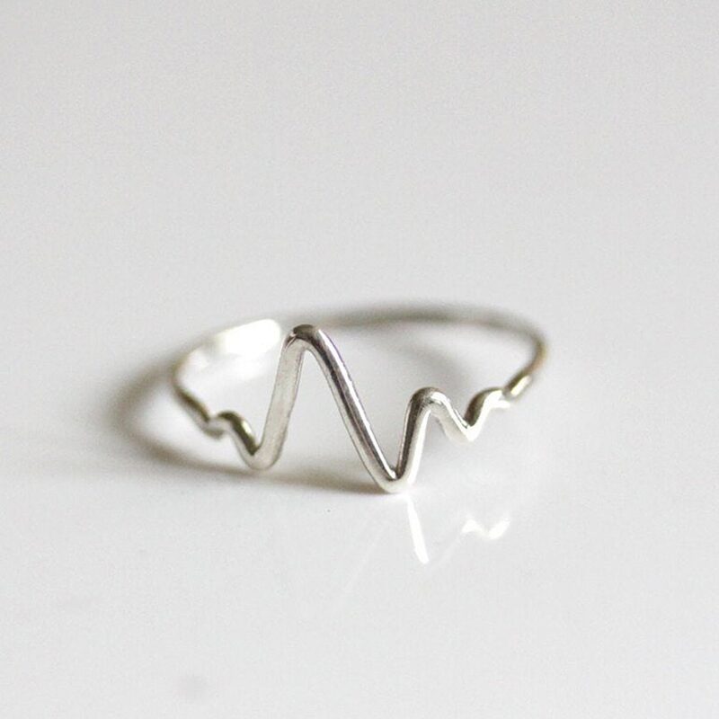 A minimalist heartbeat ring crafted in polished 925 sterling silver, featuring a continuous pulse line as the centerpiece. The delicate curve of the band mirrors the rhythm of a heart, symbolizing vitality, emotion, and connection. Perfect for nurses, doctors, or anyone who cherishes life's precious moments. Set against a clean background to highlight the reflective beauty of the metal.