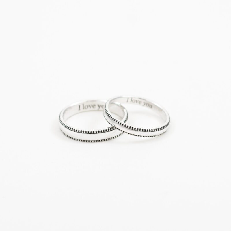 This close-up image showcases a pair of minimalist sterling silver couple rings, positioned at a gentle angle to highlight their delicate beaded edge design. The engraving "I love you" is subtly visible inside the bands, adding a personal and romantic touch. The clean white background enhances the refined silver shine, making these rings a perfect symbol of understated love and connection.