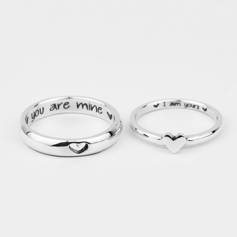 A close-up of a pair of sterling silver couple rings displayed on a white background. The man’s ring features a tiny engraved heart, while the woman’s ring showcases a raised heart at its center. Inside each band is a sweet custom engraving that reads “you are mine” and “I am yours”, adding a deeply personal touch. These matching rings beautifully symbolize connection and commitment in a minimalist and meaningful style.