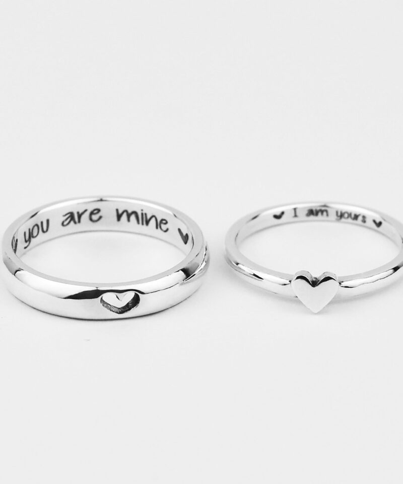 A close-up of a pair of sterling silver couple rings displayed on a white background. The man’s ring features a tiny engraved heart, while the woman’s ring showcases a raised heart at its center. Inside each band is a sweet custom engraving that reads “you are mine” and “I am yours”, adding a deeply personal touch. These matching rings beautifully symbolize connection and commitment in a minimalist and meaningful style.
