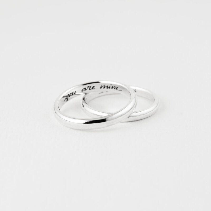 Close-up shot of two minimalist sterling silver couple rings elegantly interlocked on a white surface. One ring is engraved inside with the phrase "you are mine" in a cursive handwritten style. The mirror-like polish of the bands reflects light softly, emphasizing the sentimental value and clean craftsmanship. A perfect gift for couples seeking meaningful, custom-made jewelry with a personal touch.