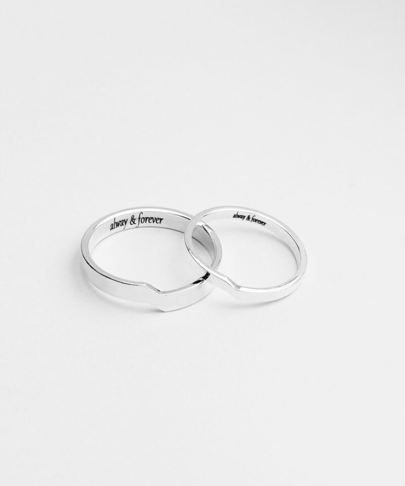 A pair of minimalist sterling silver couple rings photographed on a white background. Each ring features a subtle geometric notch that aligns perfectly when placed together, symbolizing deep connection and unity. The inner side of the rings is engraved with the phrase 