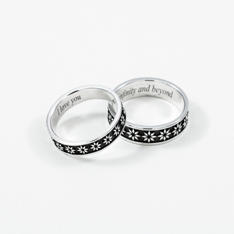 A beautifully captured pair of sterling silver couple rings featuring delicate white floral engravings on a black background. The rings are slightly overlapped to reveal the romantic custom inscriptions “I love you” and “to infinity and beyond” on the inner band. Set against a clean white backdrop, the photo highlights the polished finish and deep symbolism of eternal love and commitment between two souls.
