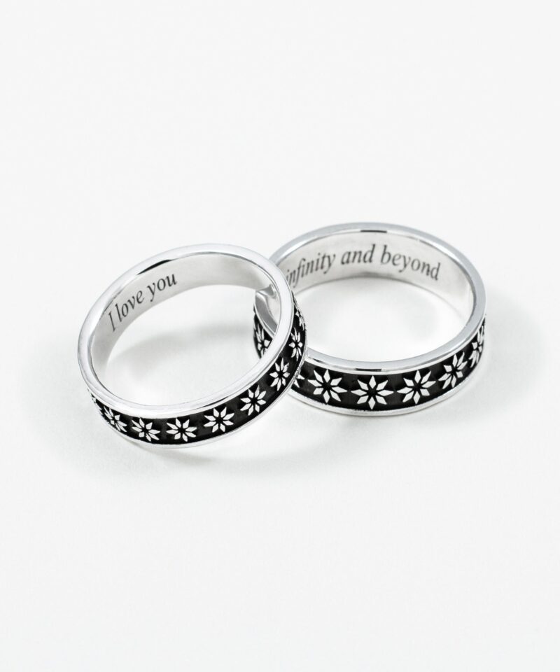 A beautifully captured pair of sterling silver couple rings featuring delicate white floral engravings on a black background. The rings are slightly overlapped to reveal the romantic custom inscriptions “I love you” and “to infinity and beyond” on the inner band. Set against a clean white backdrop, the photo highlights the polished finish and deep symbolism of eternal love and commitment between two souls.