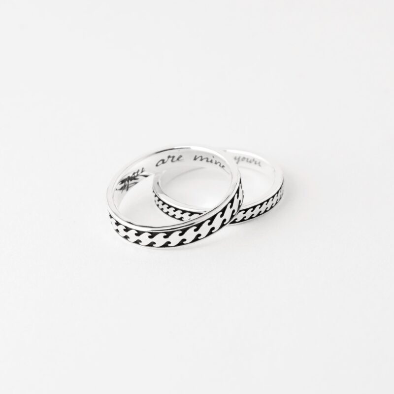 A romantic flat lay of two sterling silver Japanese wave rings resting together on a clean white background. The darkened engraving contrasts beautifully against the bright silver, highlighting the intricate movement of the wave pattern. Subtle inscriptions are visible inside the rings, offering a glimpse of personalized messages. This serene setup celebrates eternal love and the graceful flow of time shared between two hearts.