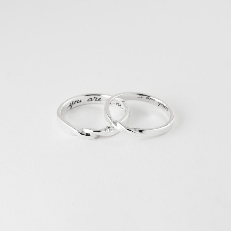Silver Mobius Twist couple rings gently overlapped on a clean white surface. The soft sculptural twist at the center adds dynamic flow to the design, while engraved messages inside personalize each piece. A timeless jewelry set that speaks of commitment and harmony.