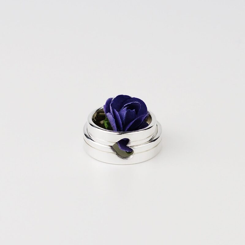 A close-up photo of two minimalist silver couple rings placed side by side with a single purple rose delicately arranged in the center. The heart-shaped cutout across both bands aligns perfectly, symbolizing unity and love when the rings are brought together. The clean white background highlights the polished sterling silver surface, giving the image a pure and romantic vibe.