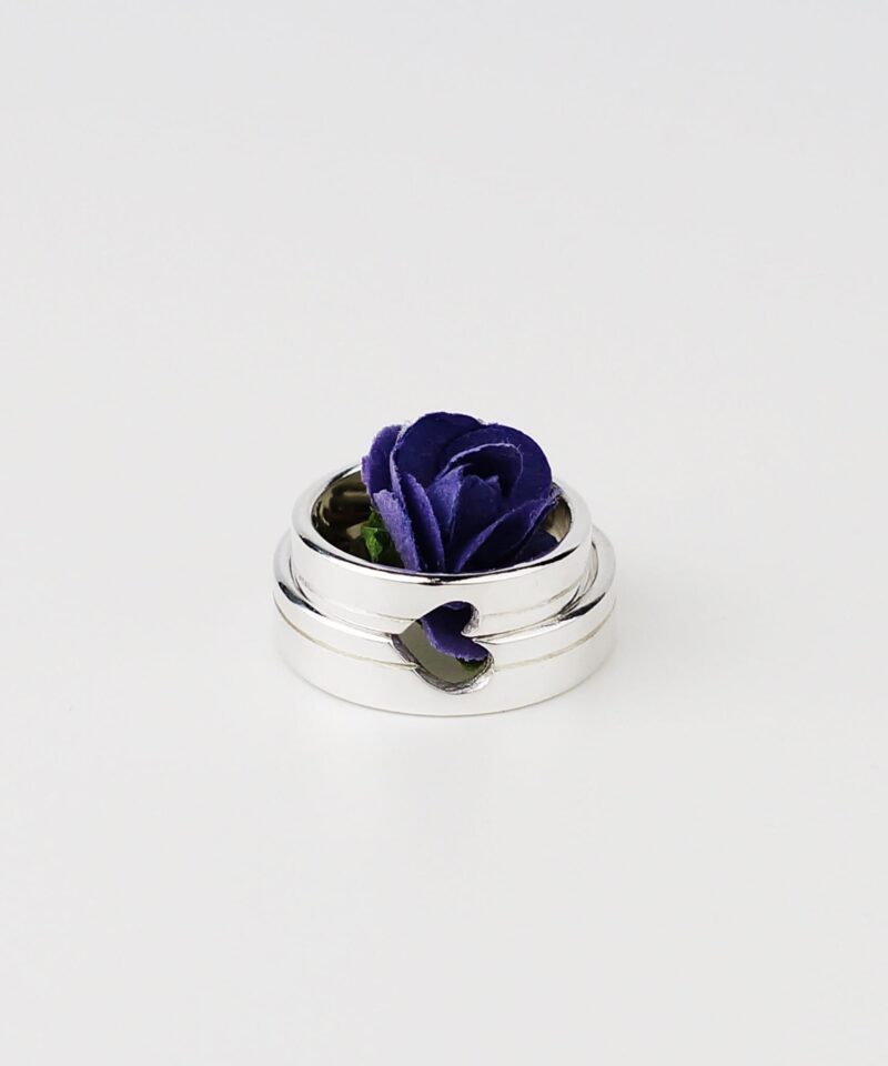 A close-up photo of two minimalist silver couple rings placed side by side with a single purple rose delicately arranged in the center. The heart-shaped cutout across both bands aligns perfectly, symbolizing unity and love when the rings are brought together. The clean white background highlights the polished sterling silver surface, giving the image a pure and romantic vibe.
