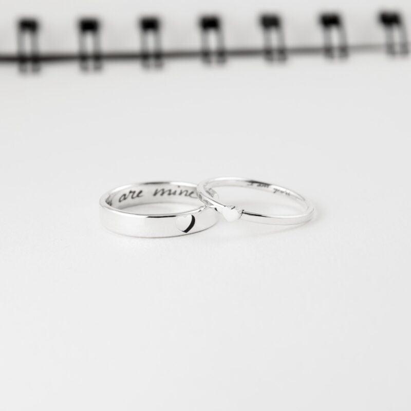 This close-up photo displays a pair of 925 sterling silver couple rings lying side by side. The men’s ring features a hollow heart, and the women’s ring has a raised heart that interlocks when placed together. Inside both rings are engraved messages in delicate script. These handcrafted rings symbolize unity and are perfect for gifting on anniversaries, weddings, or special occasions.