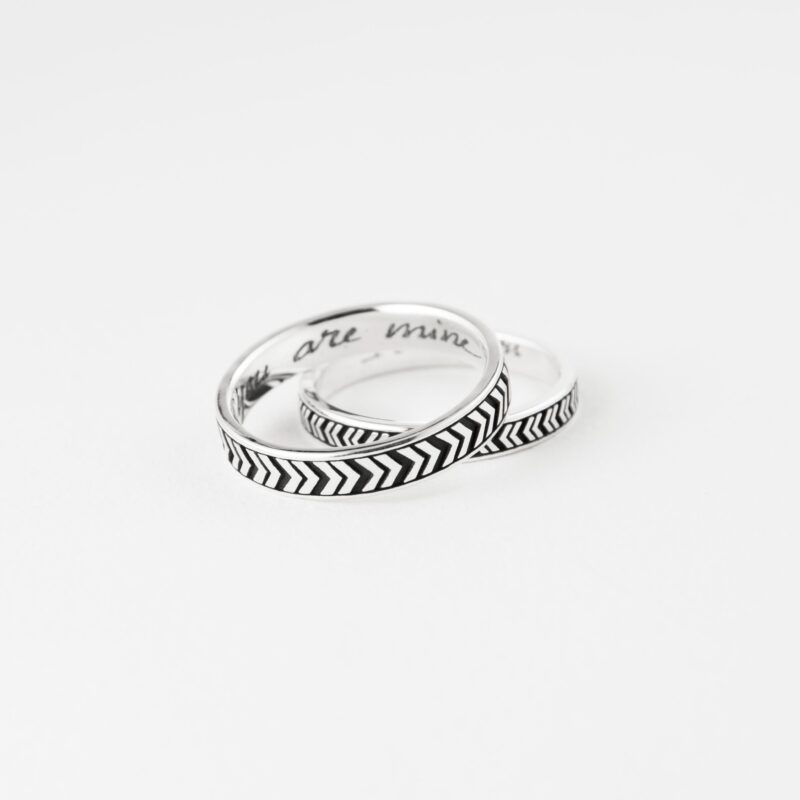 The V Arrow couple rings are gently intertwined and laid against a soft white surface, suggesting unity and closeness. The deeply etched chevron pattern adds texture and meaning to the clean silver band, symbolizing forward motion and shared direction in life. This shot captures the essence of minimalistic love jewelry—sophisticated, symbolic, and made to last through time.