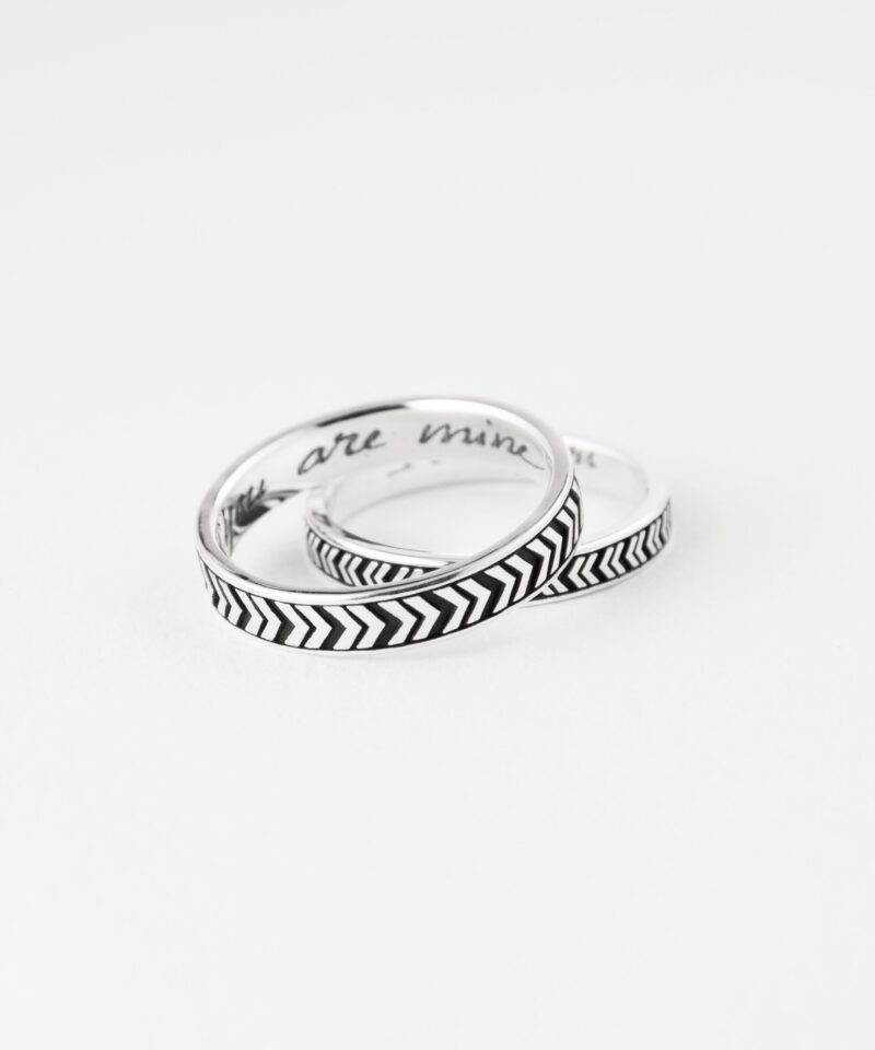 The V Arrow couple rings are gently intertwined and laid against a soft white surface, suggesting unity and closeness. The deeply etched chevron pattern adds texture and meaning to the clean silver band, symbolizing forward motion and shared direction in life. This shot captures the essence of minimalistic love jewelry—sophisticated, symbolic, and made to last through time.