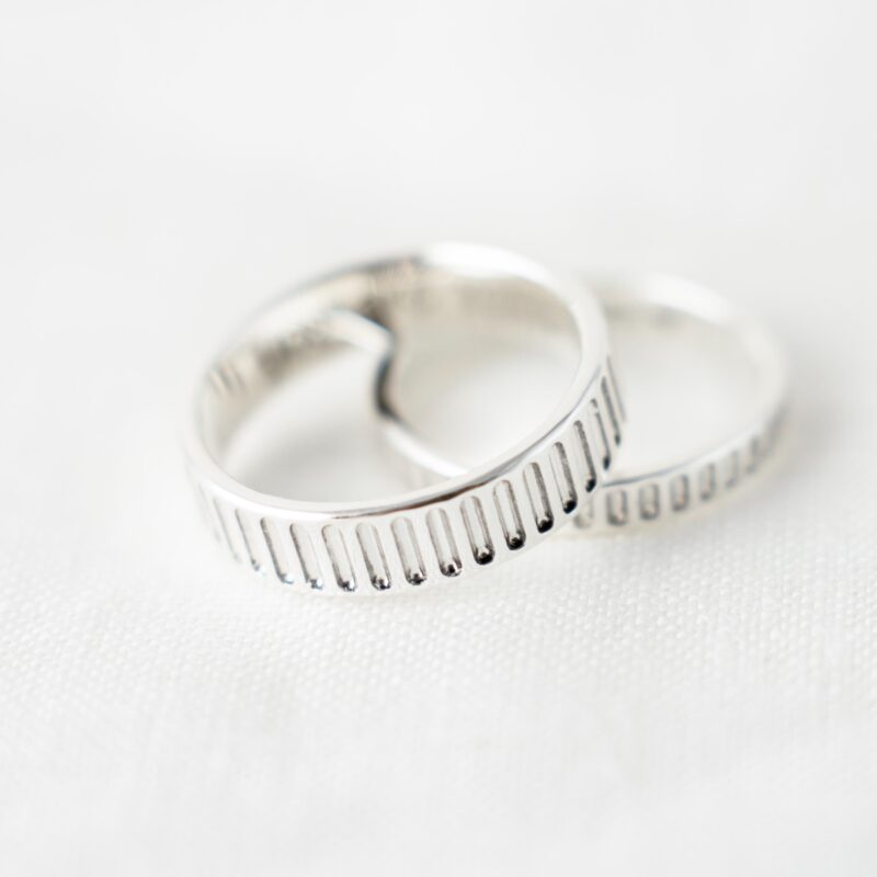 A close-up shot of a pair of 925 sterling silver couple rings placed on white fabric, each featuring a distinctive vertical line pattern engraved along the outer band. The elegant groove detailing reflects the light softly, highlighting the contrast between polish and depth. These minimalist matching rings symbolize unity and timeless design—perfect for lovers who appreciate clean lines and subtle texture in everyday jewelry.