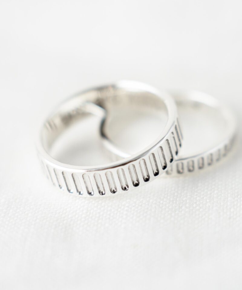 A close-up shot of a pair of 925 sterling silver couple rings placed on white fabric, each featuring a distinctive vertical line pattern engraved along the outer band. The elegant groove detailing reflects the light softly, highlighting the contrast between polish and depth. These minimalist matching rings symbolize unity and timeless design—perfect for lovers who appreciate clean lines and subtle texture in everyday jewelry.