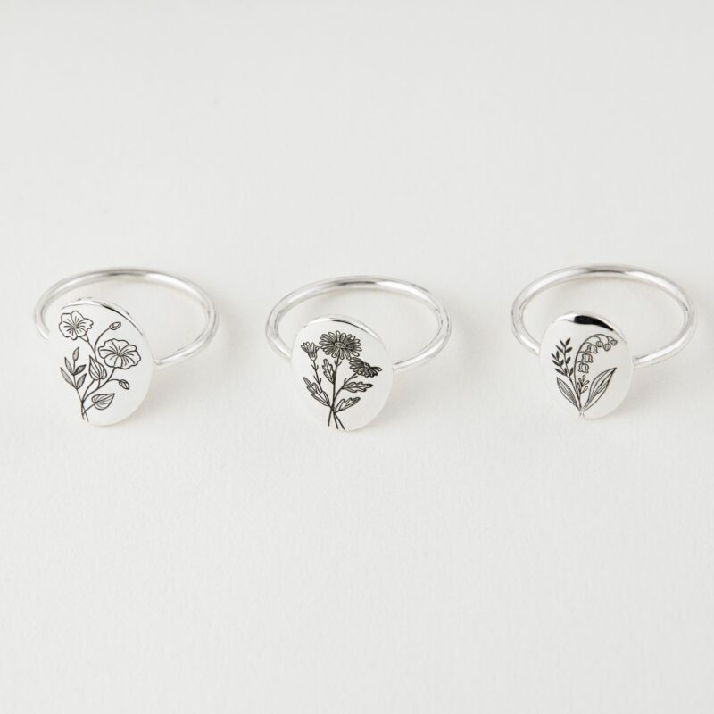 Overhead view of three silver birth flower rings arranged in a line on a clean white background, each engraved with a different minimalist botanical design. The unique floral motifs represent various birth months, perfect for personalized gifts.