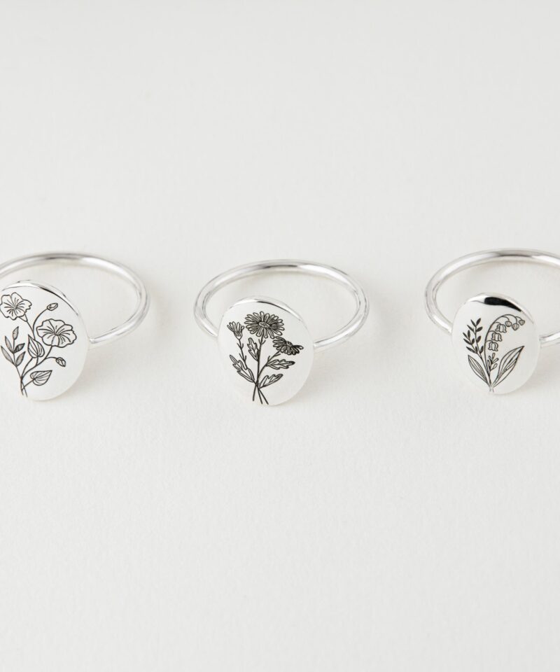 Overhead view of three silver birth flower rings arranged in a line on a clean white background, each engraved with a different minimalist botanical design. The unique floral motifs represent various birth months, perfect for personalized gifts.