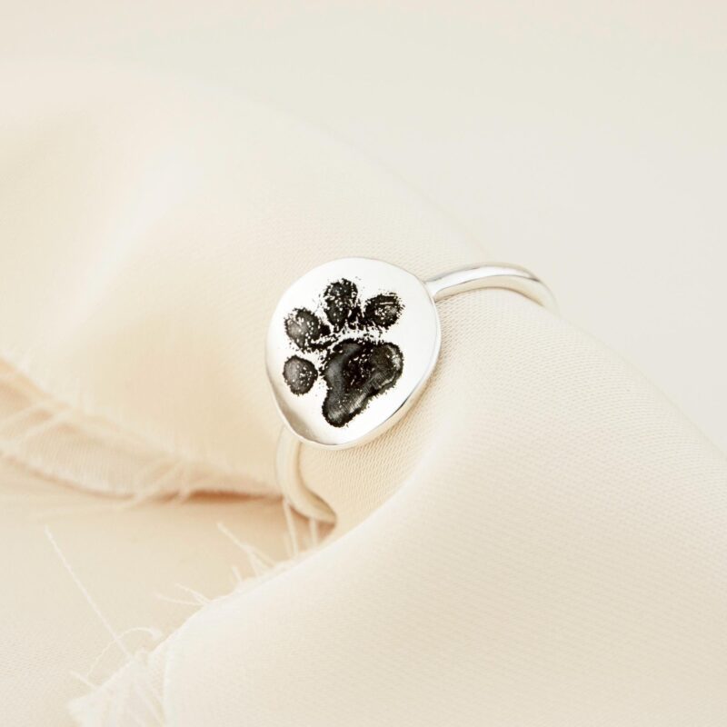 This image shows a paw print ring resting on cream-colored linen fabric, highlighting the glossy finish and natural details of the engraved pet footprint. The ring reflects a meaningful connection between pet and owner, crafted in solid sterling silver with an organic, smooth band suitable for everyday wear. A lovely keepsake for pet parents cherishing lasting bonds.