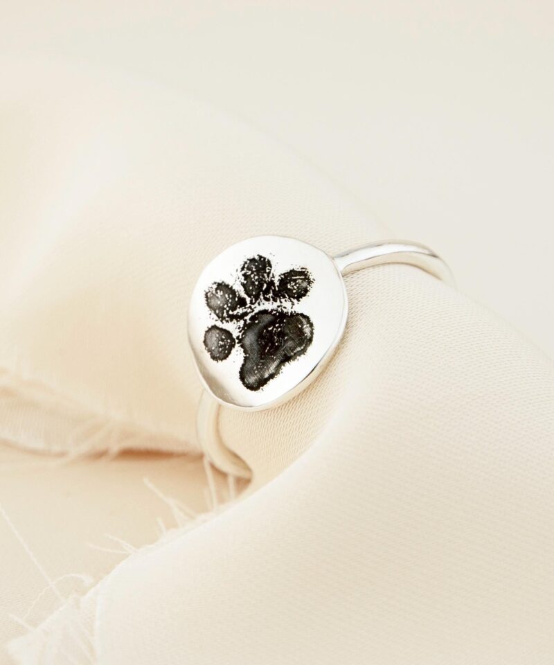 This image shows a paw print ring resting on cream-colored linen fabric, highlighting the glossy finish and natural details of the engraved pet footprint. The ring reflects a meaningful connection between pet and owner, crafted in solid sterling silver with an organic, smooth band suitable for everyday wear. A lovely keepsake for pet parents cherishing lasting bonds.