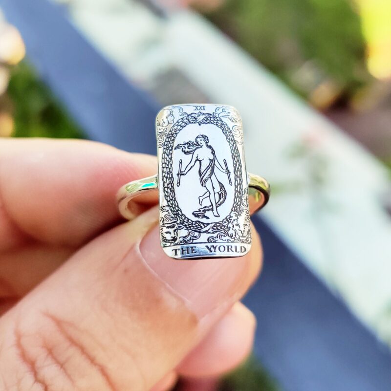 Close-up of a sterling silver rectangular ring featuring The World tarot card design from the Rider-Waite deck. The highly detailed black and white engraving shows the figure within a laurel wreath, surrounded by symbolic figures, captured in natural sunlight while held between fingers.