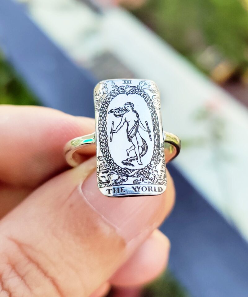 Close-up of a sterling silver rectangular ring featuring The World tarot card design from the Rider-Waite deck. The highly detailed black and white engraving shows the figure within a laurel wreath, surrounded by symbolic figures, captured in natural sunlight while held between fingers.