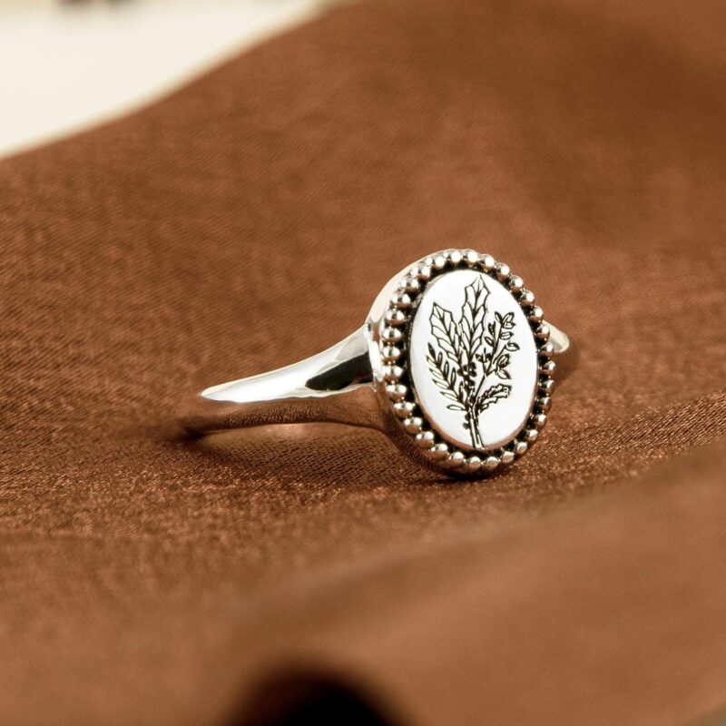 Minimalist botanical silver ring photographed diagonally on a warm fabric surface. The engraved birth flower illustration is clean and detailed, enclosed by tiny silver beads along the edge of the signet-style oval face. Crafted in 925 sterling silver, this ring makes a unique personalized gift for birthday, floral lovers, or those who appreciate nature-inspired talismans.
