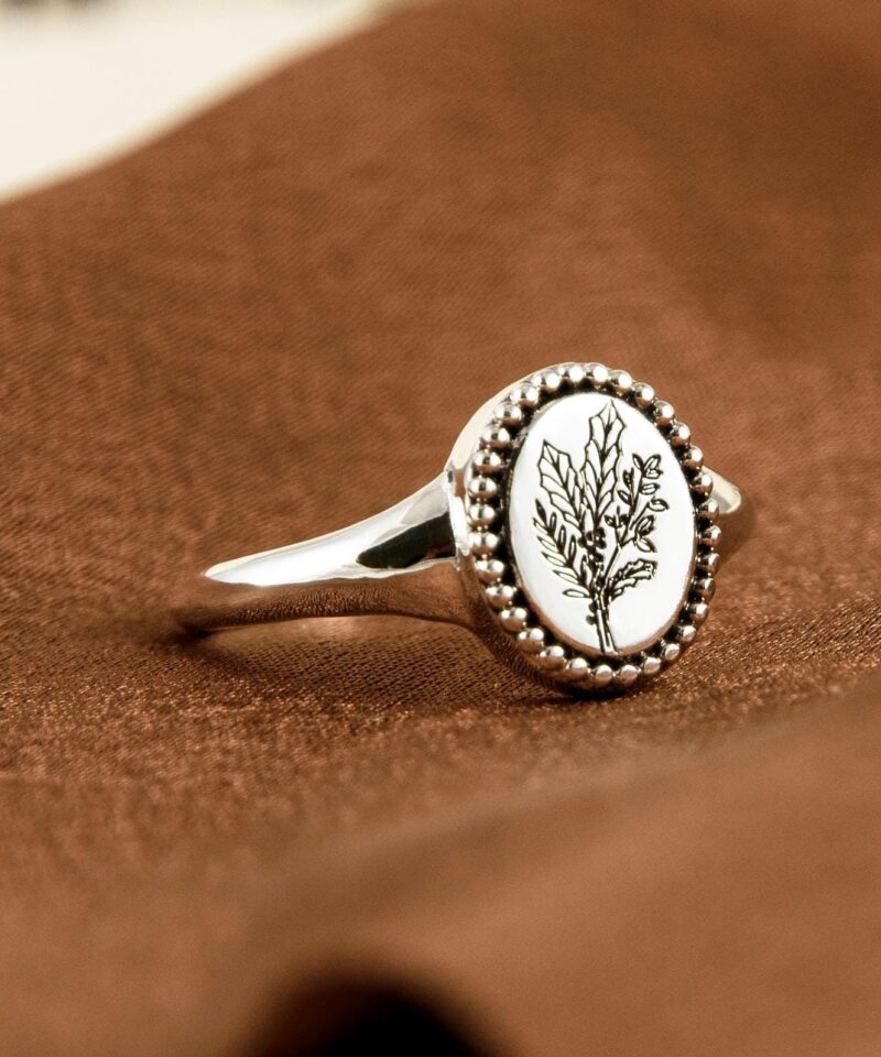 Minimalist botanical silver ring photographed diagonally on a warm fabric surface. The engraved birth flower illustration is clean and detailed, enclosed by tiny silver beads along the edge of the signet-style oval face. Crafted in 925 sterling silver, this ring makes a unique personalized gift for birthday, floral lovers, or those who appreciate nature-inspired talismans.