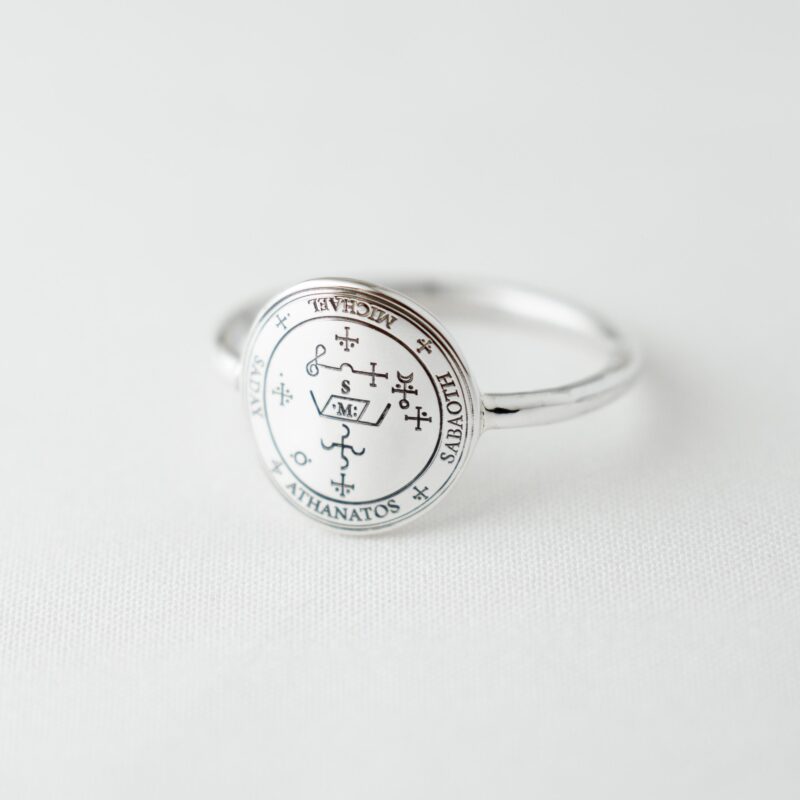 Close-up image of a silver signet ring engraved with the Sigil of Archangel Michael, placed on white fabric. The elegant black line engraving features sacred names such as Michael, Sabaoth, and Athanatos. The polished 925 sterling silver surface reflects soft light, highlighting the minimalist yet spiritual appeal of this protective talisman jewelry.