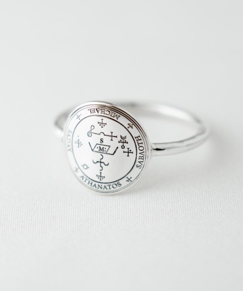 Close-up image of a silver signet ring engraved with the Sigil of Archangel Michael, placed on white fabric. The elegant black line engraving features sacred names such as Michael, Sabaoth, and Athanatos. The polished 925 sterling silver surface reflects soft light, highlighting the minimalist yet spiritual appeal of this protective talisman jewelry.