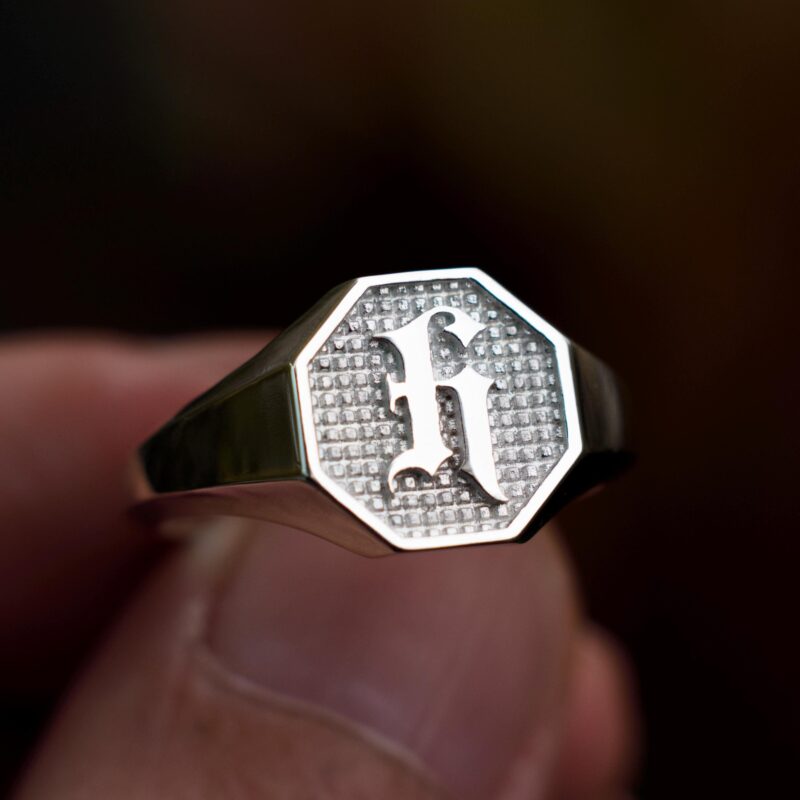Handheld shot of an initial signet ring in polished sterling silver, showing its bold octagonal face with a gothic "H" rising from a textured silver field. Designed as a modern heirloom piece, this ring is ideal for gifting to fathers, sons, or partners. Its balance between vintage masculinity and clean lines makes it a standout among custom engraved silver rings, with spiritual, personal, and historical appeal.