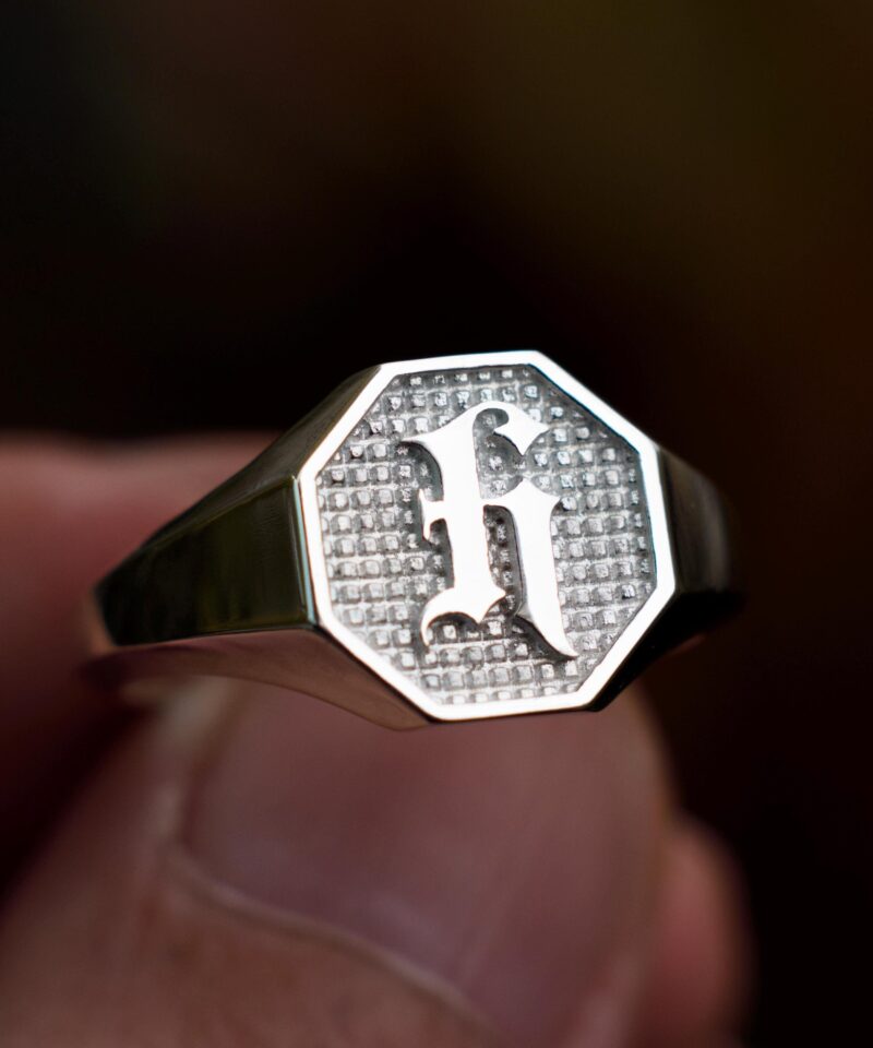 Handheld shot of an initial signet ring in polished sterling silver, showing its bold octagonal face with a gothic "H" rising from a textured silver field. Designed as a modern heirloom piece, this ring is ideal for gifting to fathers, sons, or partners. Its balance between vintage masculinity and clean lines makes it a standout among custom engraved silver rings, with spiritual, personal, and historical appeal.